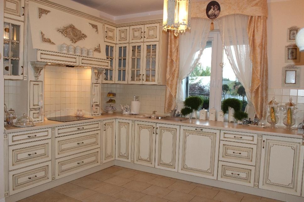 Kremowa kuchnia z frezowanymi frontami, szklanymi witrynami i dekoracyjnym okapem, uzupełniona oświetleniem podszafkowym i ozdobnymi zasłonami.