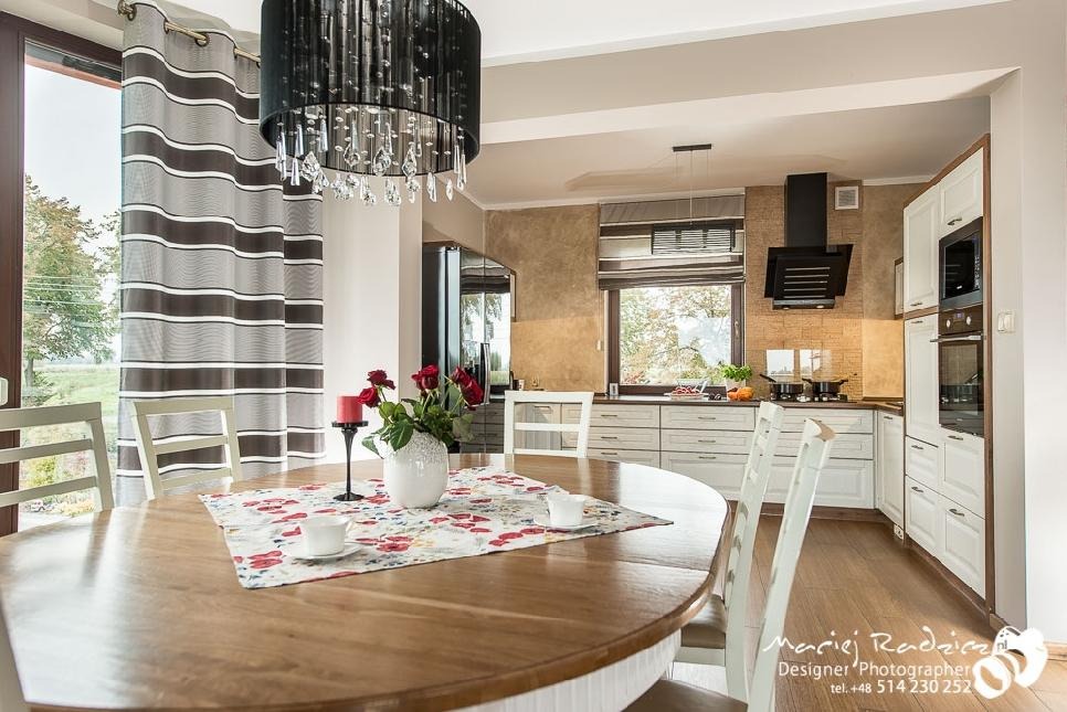 Jasna kuchnia z białymi szafkami, czarnym okapem i brązowymi akcentami, widok zza okrągłego stołu z kwiatami i zasłonami w paski.