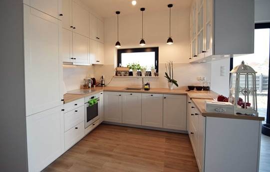 Jasna kuchnia w stylu skandynawskim z drewnianymi blatami, czarnymi uchwytami i trzema wiszącymi lampami nad oknem z ziołami.