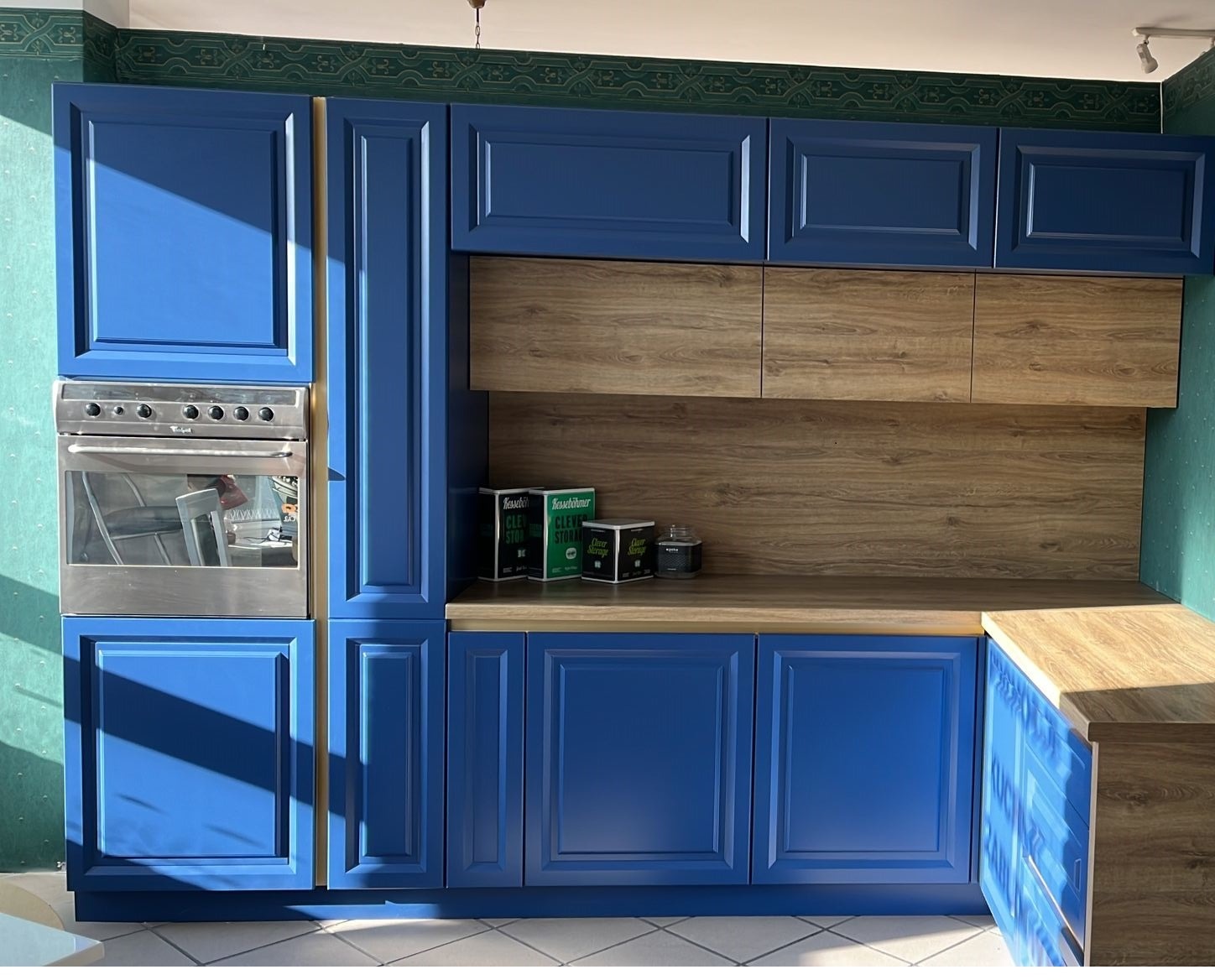 Niebieska zabudowa kuchenna z piekarnikiem, drewnianym blatem i dekoracyjną tapetą w tle.