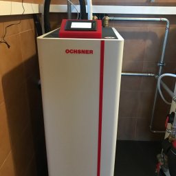 POMPY CIEPŁA.COM.PL SPÓŁKA Z OGRANICZONĄ ODPOWIEDZIALNOŚCIĄ - Biała jednostka grzewcza Ochsner z czerwonymi akcentami, zamontowana w pomieszczeniu z brązowymi płytkami, widoczne rury instalacyjne i przewody z izolacją termiczną.