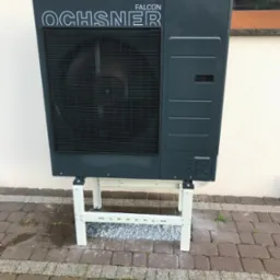 Szara jednostka zewnętrzna z wentylatorem marki Ochsner Falcon, umieszczona na białym metalowym stelażu, stoi na kostce brukowej przed jasną ścianą budynku.