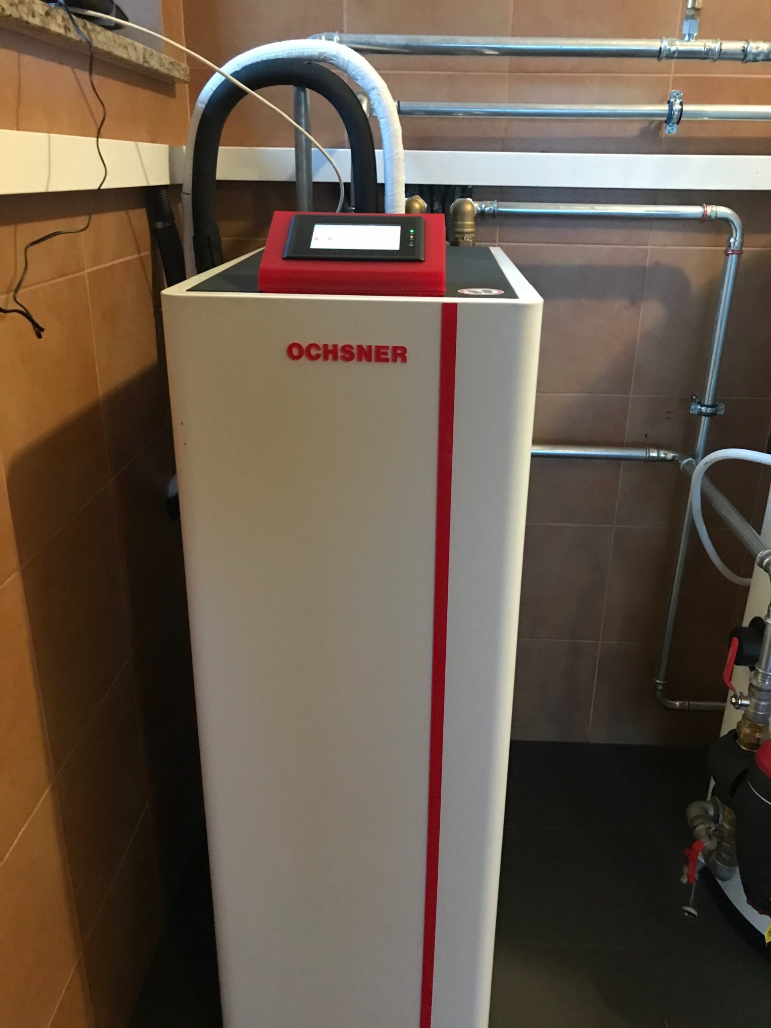 Biała jednostka grzewcza Ochsner z czerwonymi akcentami, zamontowana w pomieszczeniu z brązowymi płytkami, widoczne rury instalacyjne i przewody z izolacją termiczną.