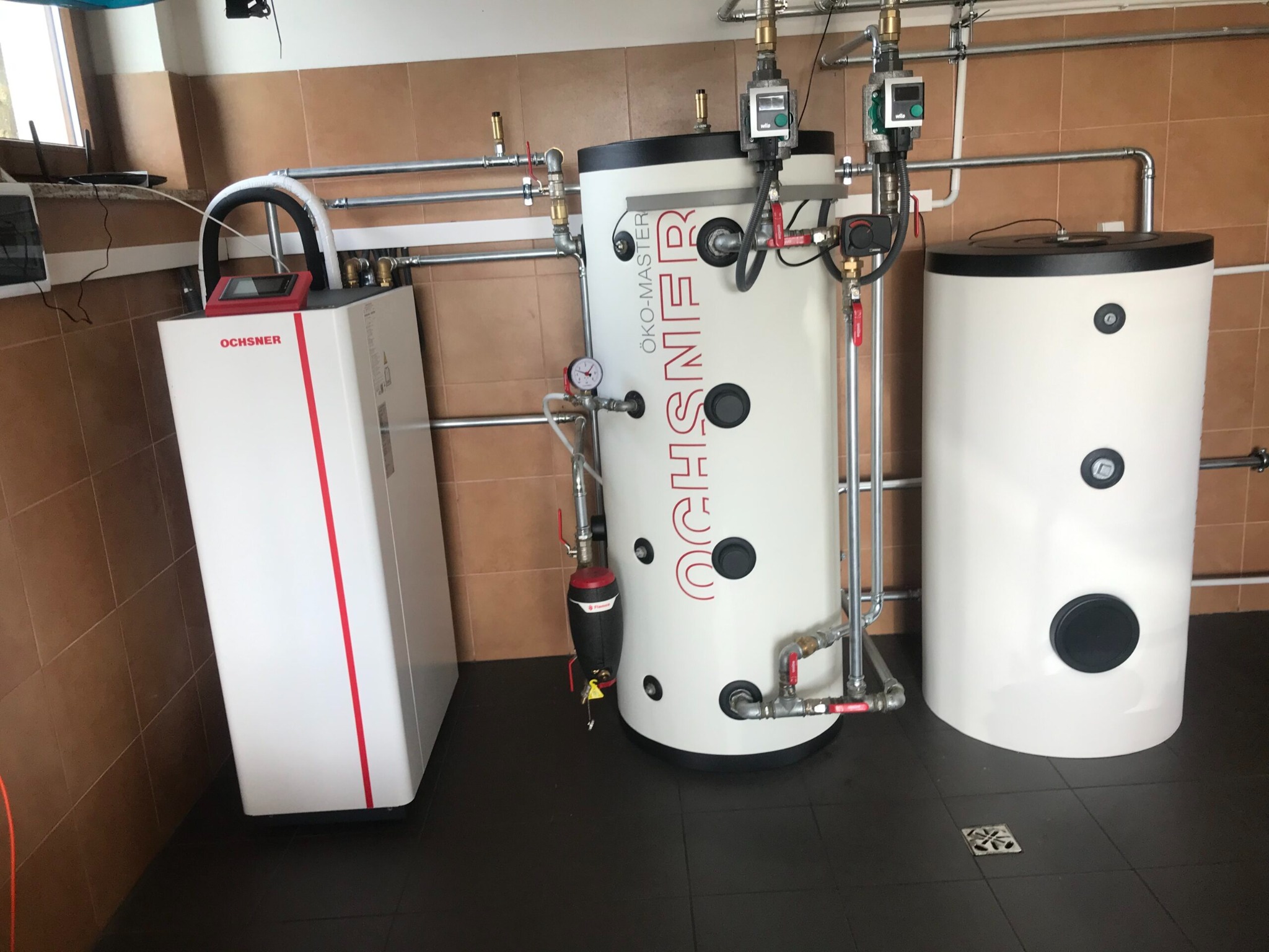 Instalacja grzewcza z trzema zbiornikami, w tym jeden z napisem 'ÖKÖ-MASTER' i drugi z napisem 'OCHSNER', podłączona rurami w pomieszczeniu z brązowymi kafelkami na ścianach i ciemną podłogą.