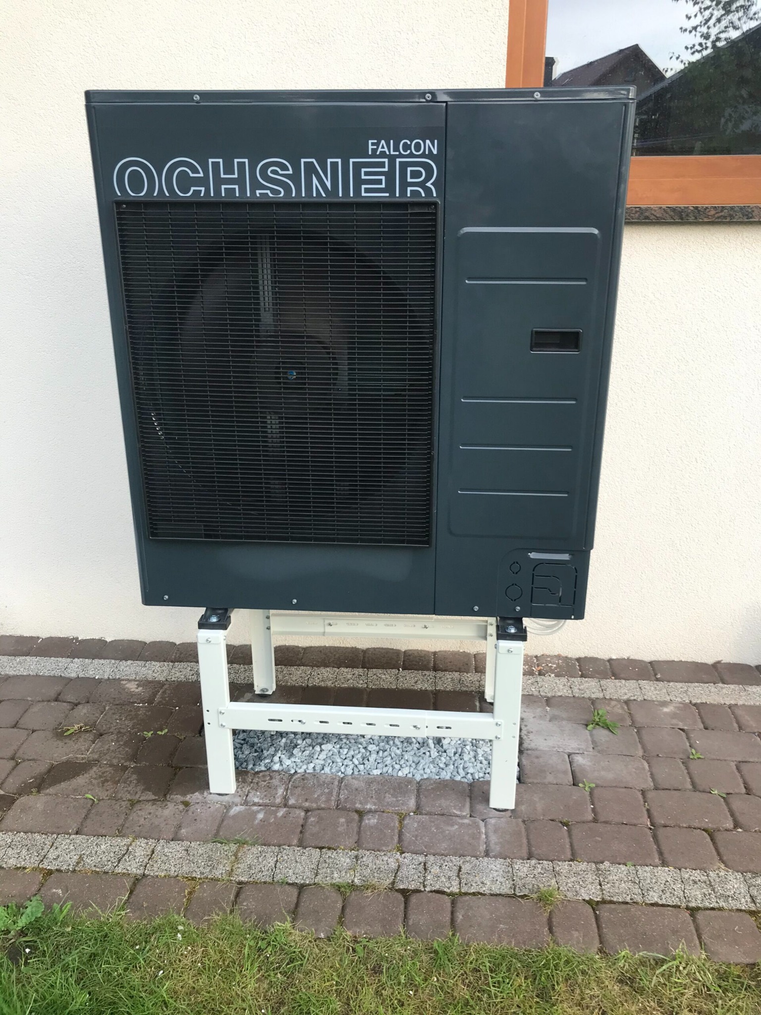 Szara jednostka zewnętrzna z wentylatorem marki Ochsner Falcon, umieszczona na białym metalowym stelażu, stoi na kostce brukowej przed jasną ścianą budynku.