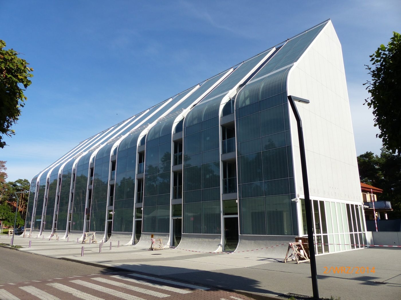 Nowoczesny budynek z przeszkloną fasadą i skośnym dachem, widoczny z perspektywy ulicy z przejściem dla pieszych, barierki ochronne na chodniku.