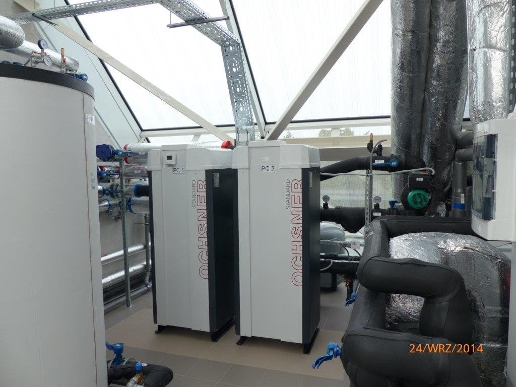 Dwie jednostki grzewcze Ochsner Standard PC1 i PC2 w kotłowni z przeszklonym dachem, widoczne rury i izolacje, manometry oraz elementy instalacji.