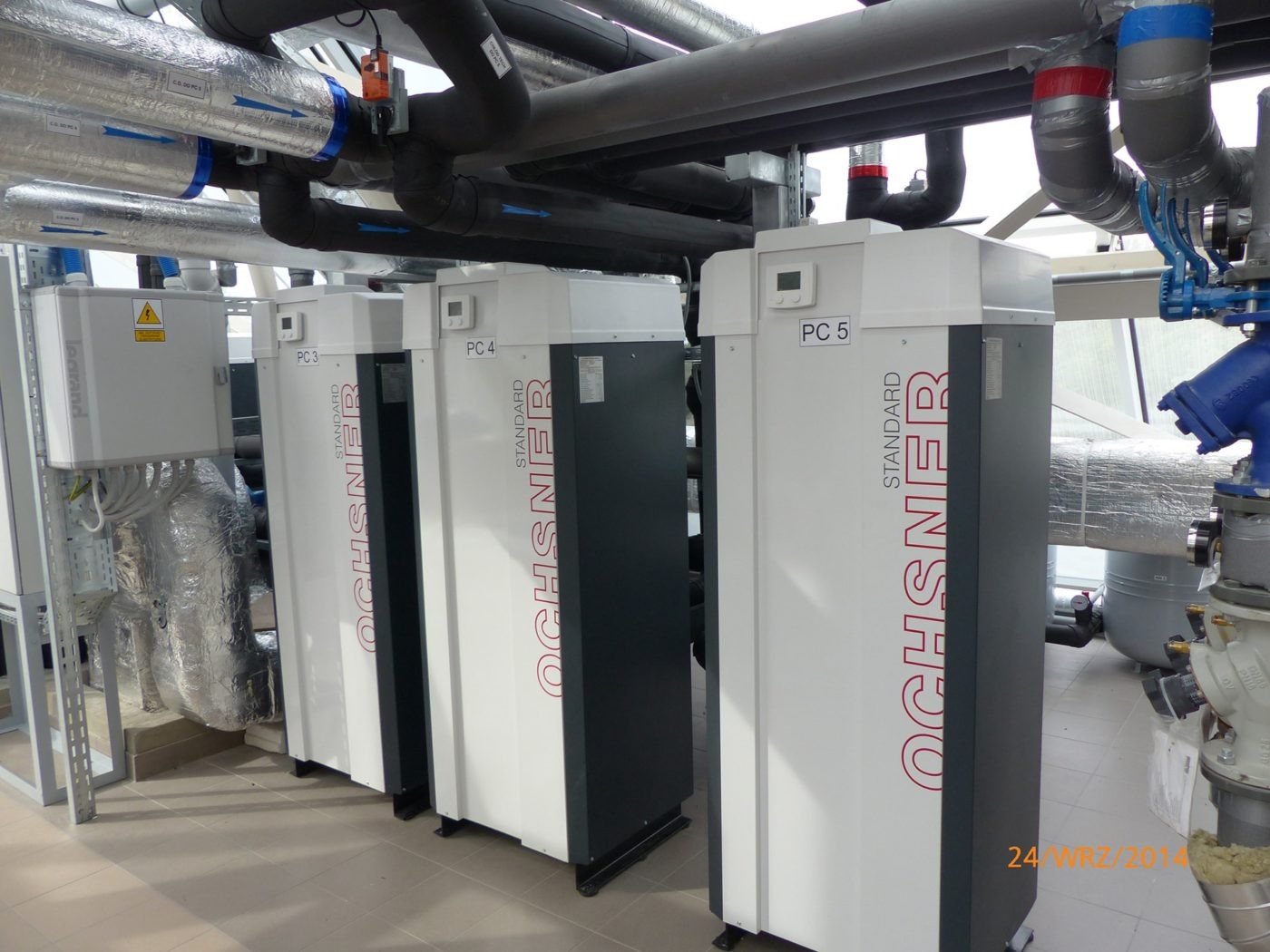 Rząd trzech jednostek grzewczych Ochsner PC 3, PC 4 i PC 5 w kotłowni, z widocznymi rurami instalacji grzewczej nad nimi.