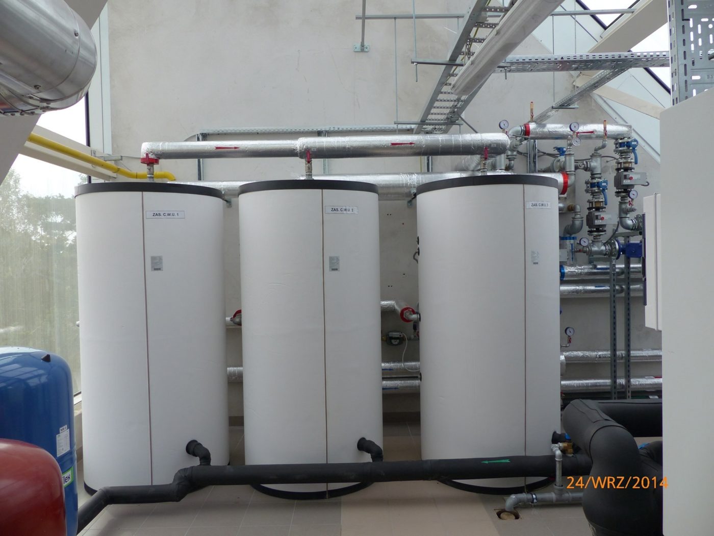 Trzy białe, cylindryczne zbiorniki z czarnymi elementami, oznaczone jako ZAS C.W.U 1, 2 i 7, połączone rurami, w jasnym pomieszczeniu z widocznymi instalacjami i datą 24/WRZ/2014 w prawym dolnym rogu.