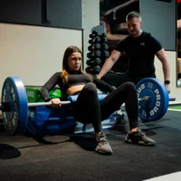 Kobieta wykonuje ćwiczenie hip thrust z obciążeniem pod nadzorem trenera w siłowni; niebieskie obciążniki PROUD, czarne ubranie sportowe.