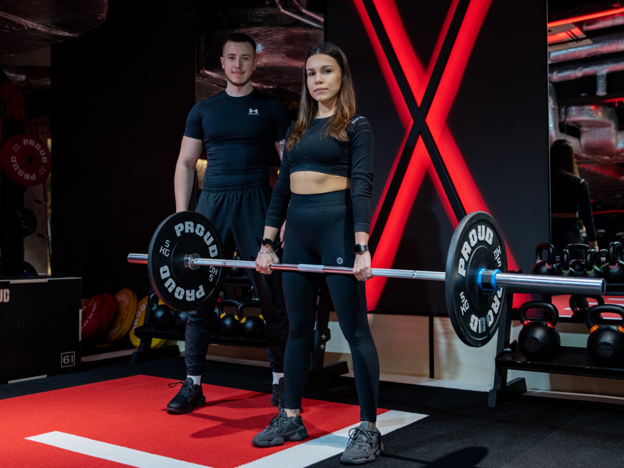 Kobieta i mężczyzna w czarnych strojach sportowych trzymają sztangę z obciążeniem w siłowni z czerwonymi akcentami i lustrem