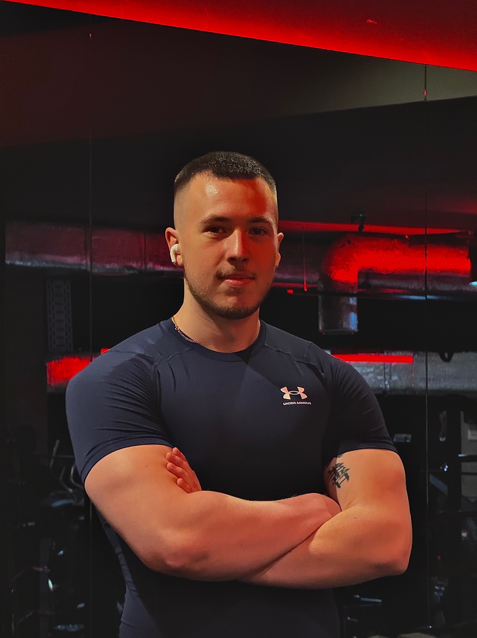 Młody mężczyzna w sportowej koszulce Under Armour z założonymi rękami na tle siłowni z czerwonym oświetleniem