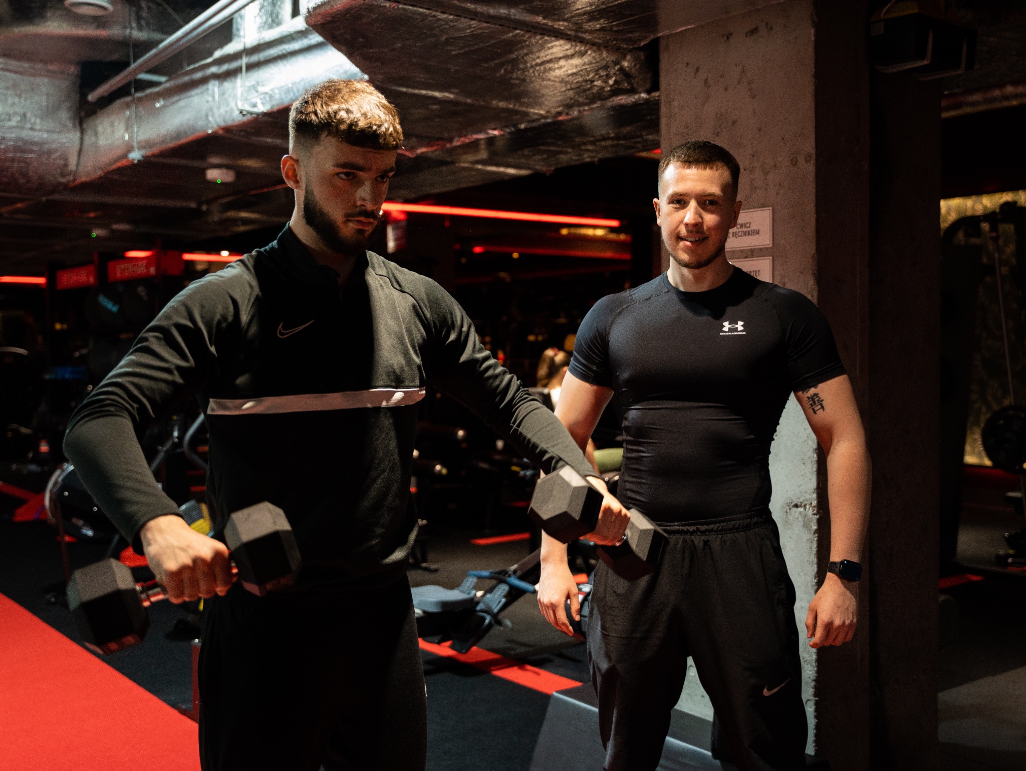 Dwóch mężczyzn w czarnych strojach sportowych pozuje z hantlami w siłowni z czerwonym oświetleniem akcentującym. Jeden z nich ma tatuaż na ramieniu.