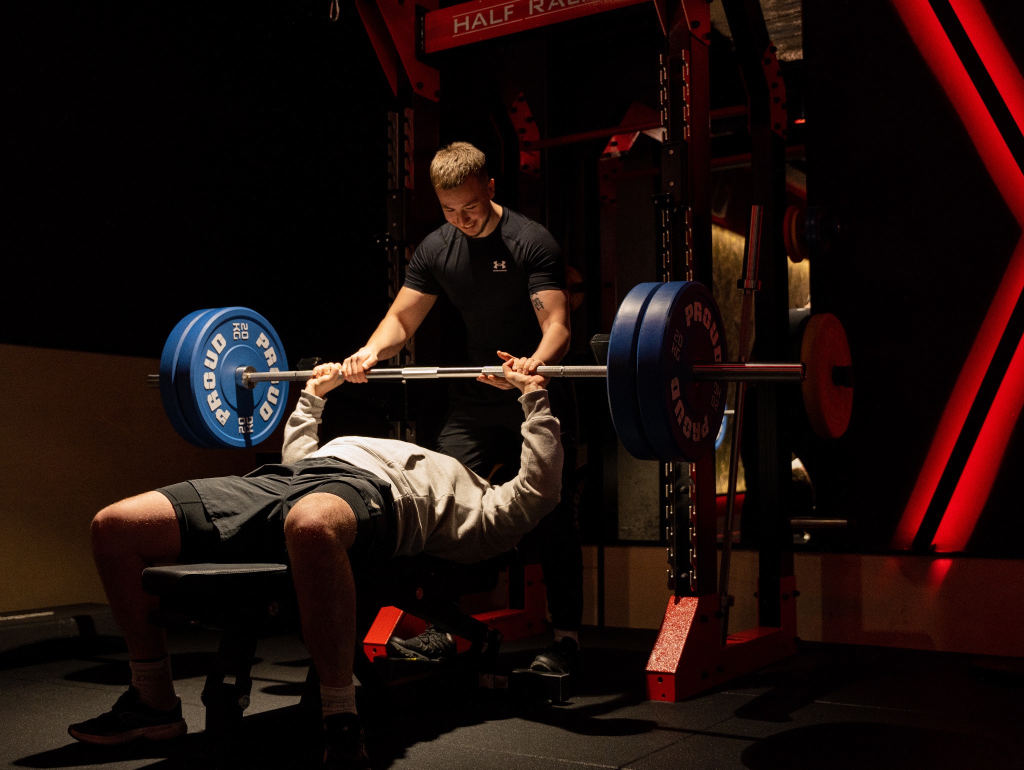 Trener personalny asekuruje mężczyznę podczas wyciskania sztangi na ławce w siłowni z czerwonymi akcentami.