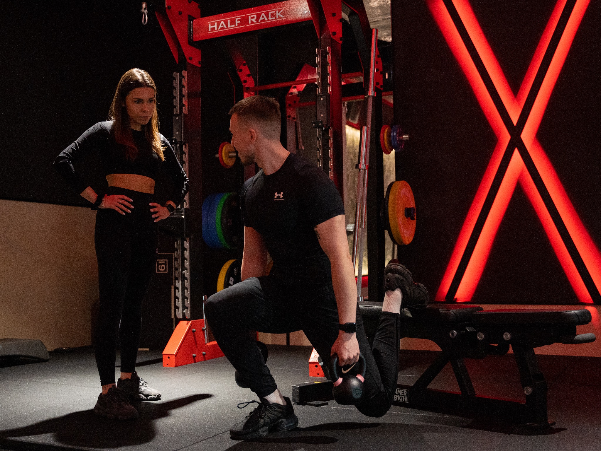 Mężczyzna wykonuje przysiad bułgarski z kettlebellem w siłowni, pod nadzorem kobiety, w tle czerwone neonowe X