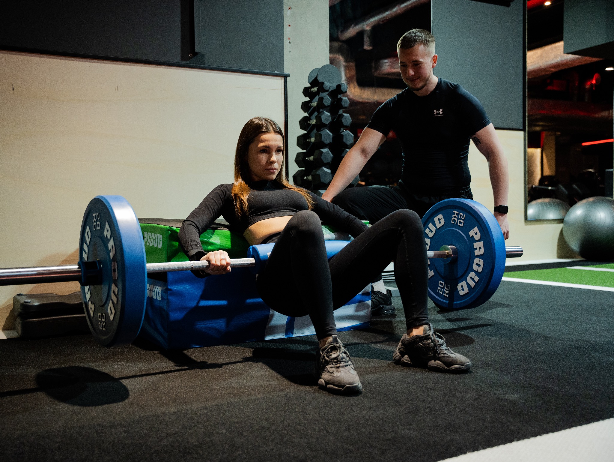 Kobieta wykonuje ćwiczenie hip thrust z obciążeniem pod nadzorem trenera w siłowni; niebieskie obciążniki PROUD, czarne ubranie sportowe.