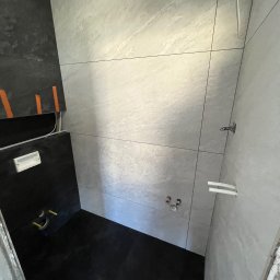 GLAZ-TAR DANIEL TARNOWSKI - Wykończona łazienka z czarno-szarą glazurą. Widoczne rury, instalacje i fragment niedokończonej ściany. Minimalistyczny design z akcentami surowego betonu.