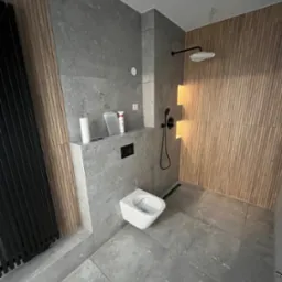 Nowoczesna łazienka z szarymi płytkami i drewnianymi lamelami. Wisząca toaleta, czarna armatura prysznicowa. Minimalistyczny design z akcentem na naturalne materiały.