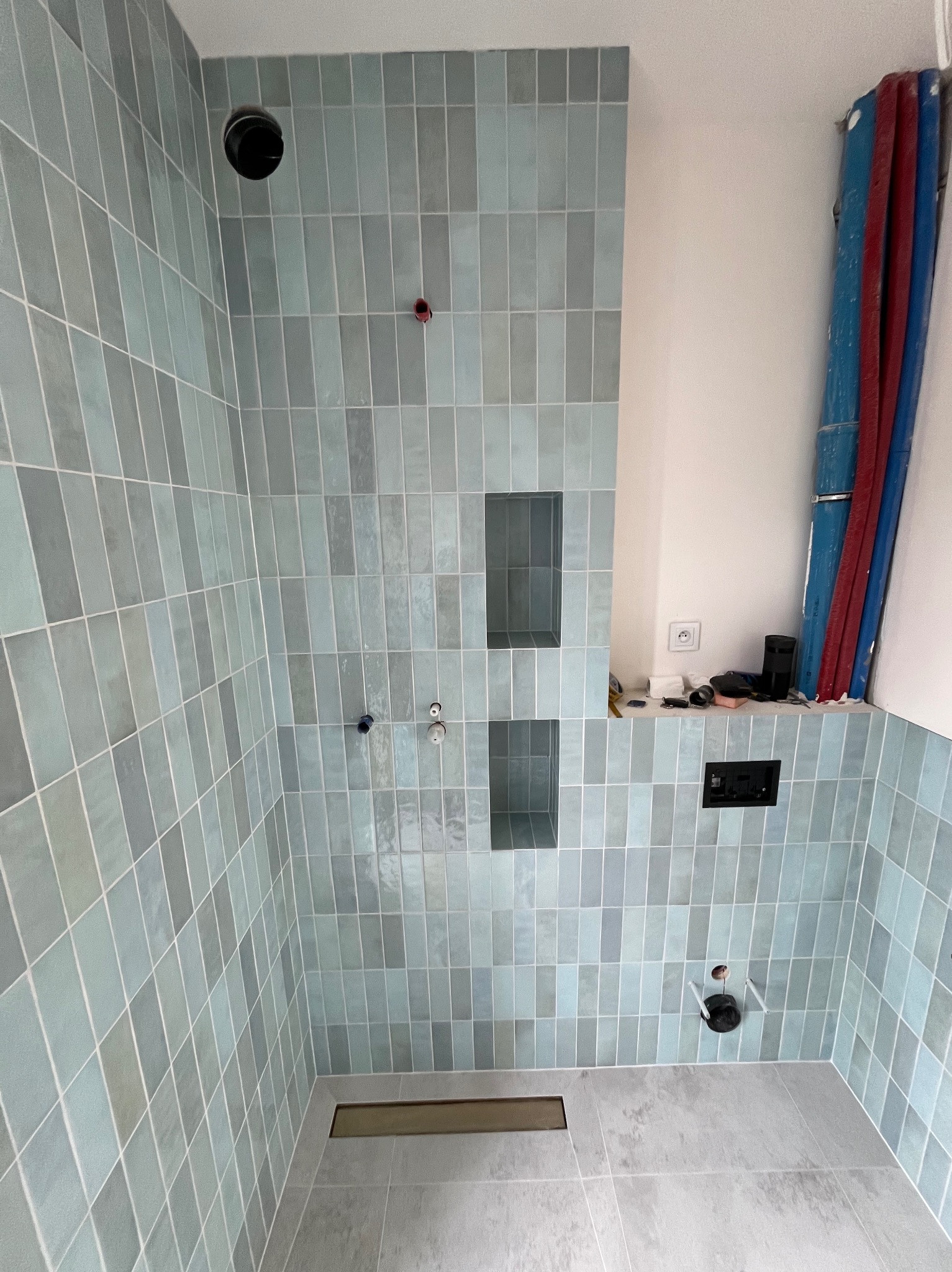 Łazienka w trakcie wykańczania: turkusowe, pionowo ułożone płytki, wnęki w ścianie, odpływ liniowy w podłodze. Instalacja hydrauliczna widoczna.