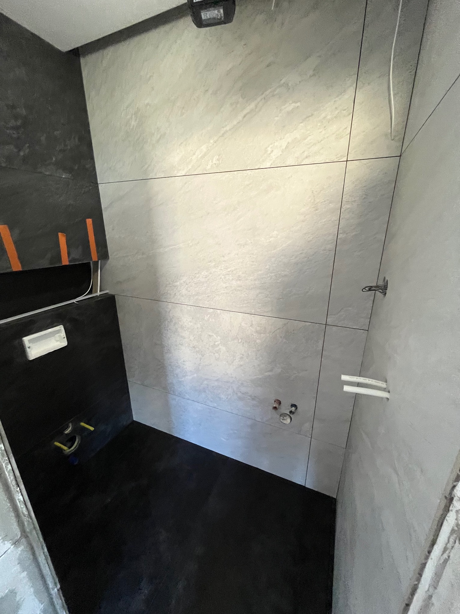 Wykończona łazienka z czarno-szarą glazurą. Widoczne rury, instalacje i fragment niedokończonej ściany. Minimalistyczny design z akcentami surowego betonu.