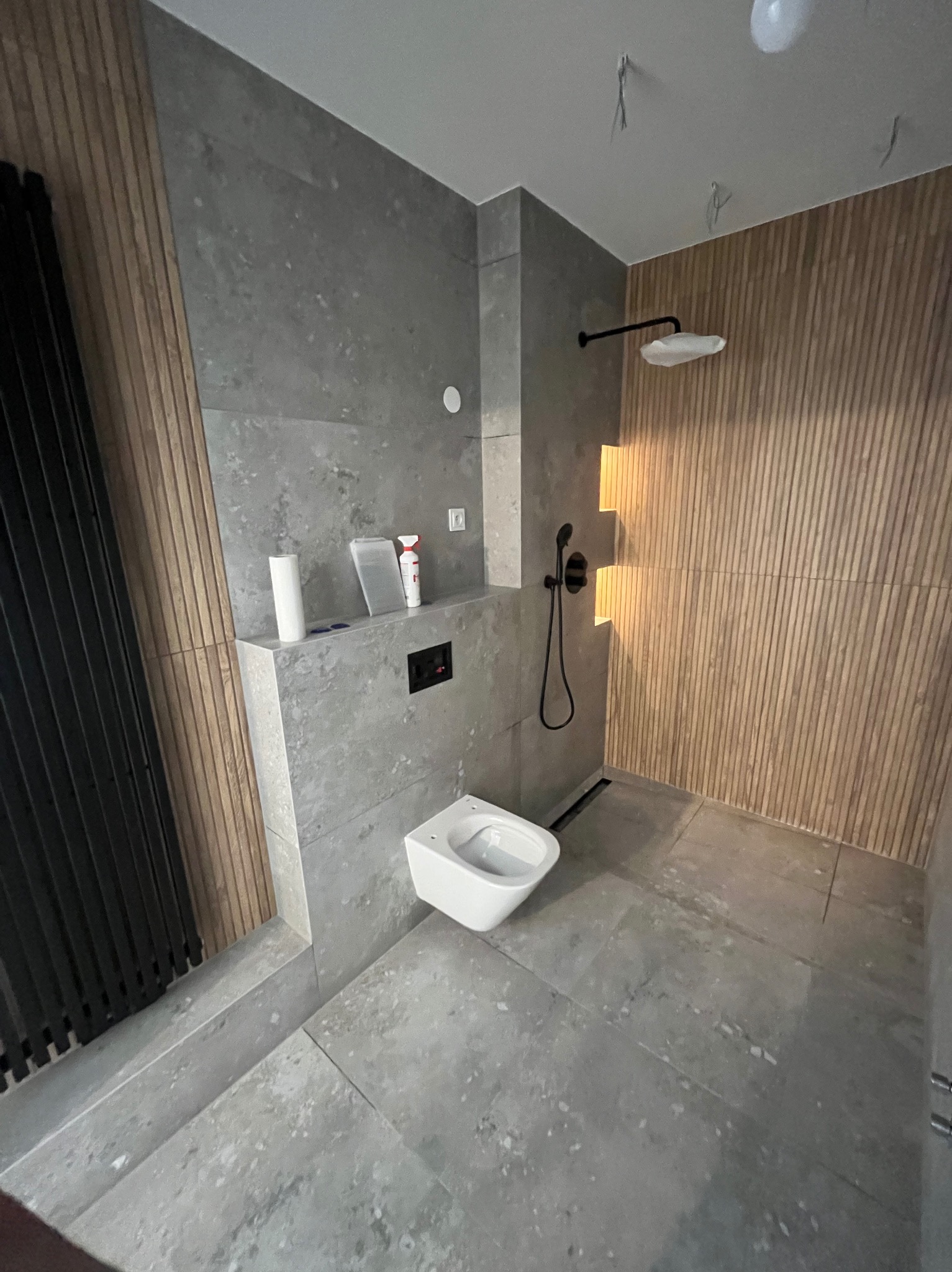 Nowoczesna łazienka z szarymi płytkami i drewnianymi lamelami. Wisząca toaleta, czarna armatura prysznicowa. Minimalistyczny design z akcentem na naturalne materiały.