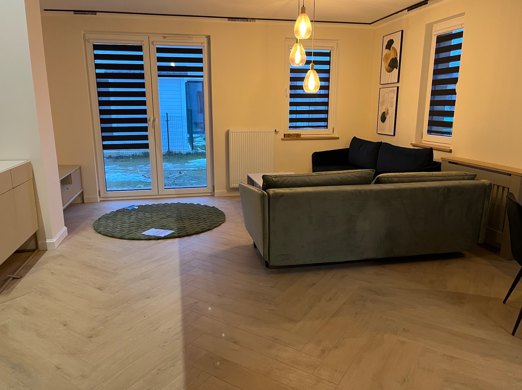 Nowoczesny salon z parkietem ułożonym w jodełkę, zieloną sofą i oknami z roletami dzień-noc. Nad sofą wiszą trzy lampy. W tle widoczny ogród przez okno.