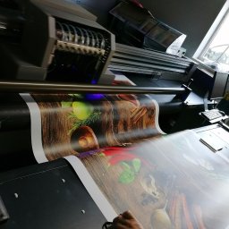 ADMEDIA - Wielkoformatowa drukarka atramentowa w trakcie drukowania grafiki przedstawiającej kompozycję kulinarną z przyprawami i ziołami na drewnianym tle.