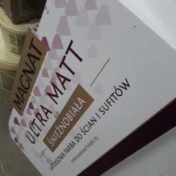 ADMEDIA - Stos tablic reklamowych farb Magnat Ultra Matt Śnieżnobiała, widoczne opakowania farb w tle.