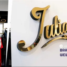Złoty, przestrzenny napis 'Julia' z czarnym obrysem, zamontowany na białej ścianie, widoczny również w sklepie odzieżowym nad wieszakami z ubraniami.