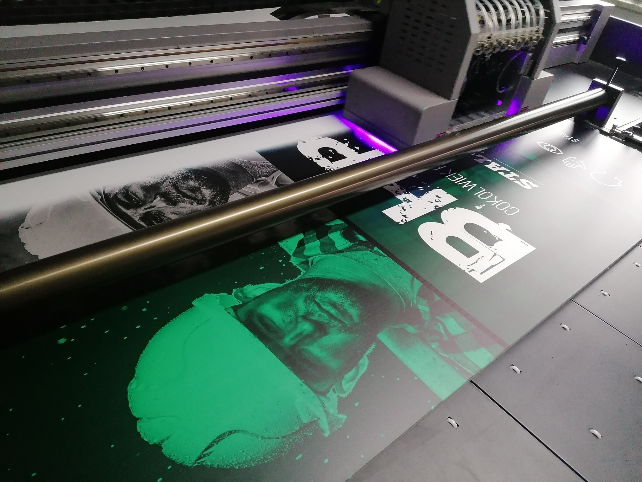 Drukarka UV w trakcie nanoszenia grafiki z portretem mężczyzny na ciemne podłoże. Widoczne głowice drukujące i element mocujący materiał.