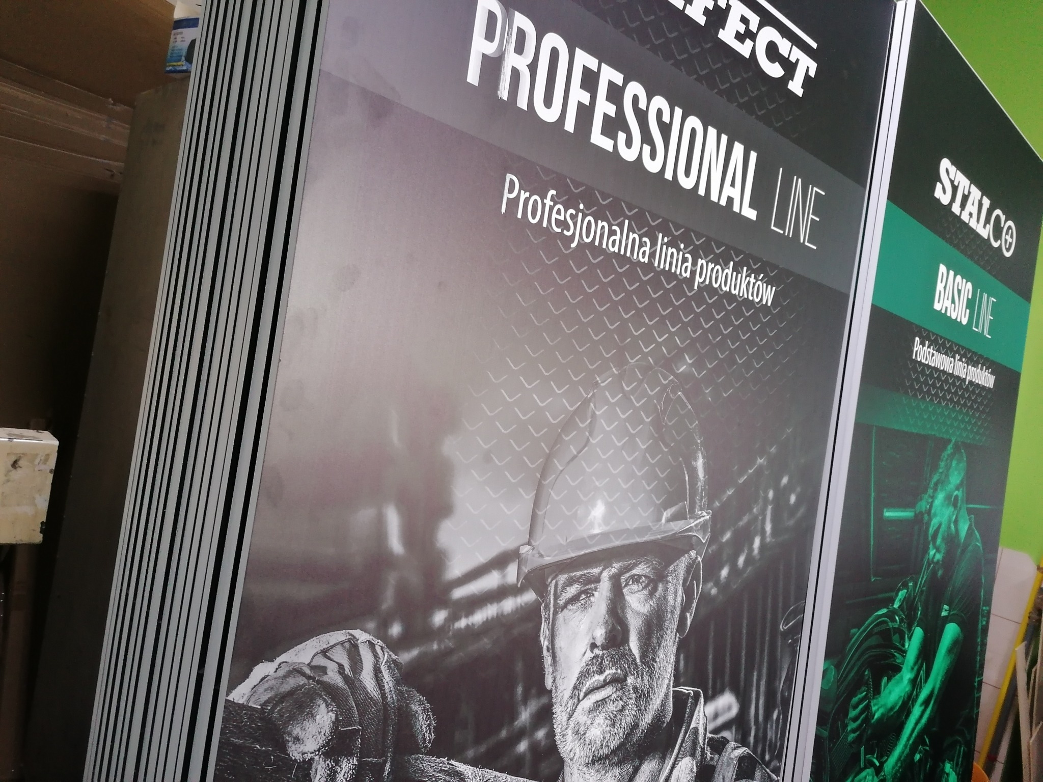 Stos wydrukowanych plakatów reklamowych z grafiką przedstawiającą robotnika w kasku i rękawicach, promujących profesjonalną linię produktów.