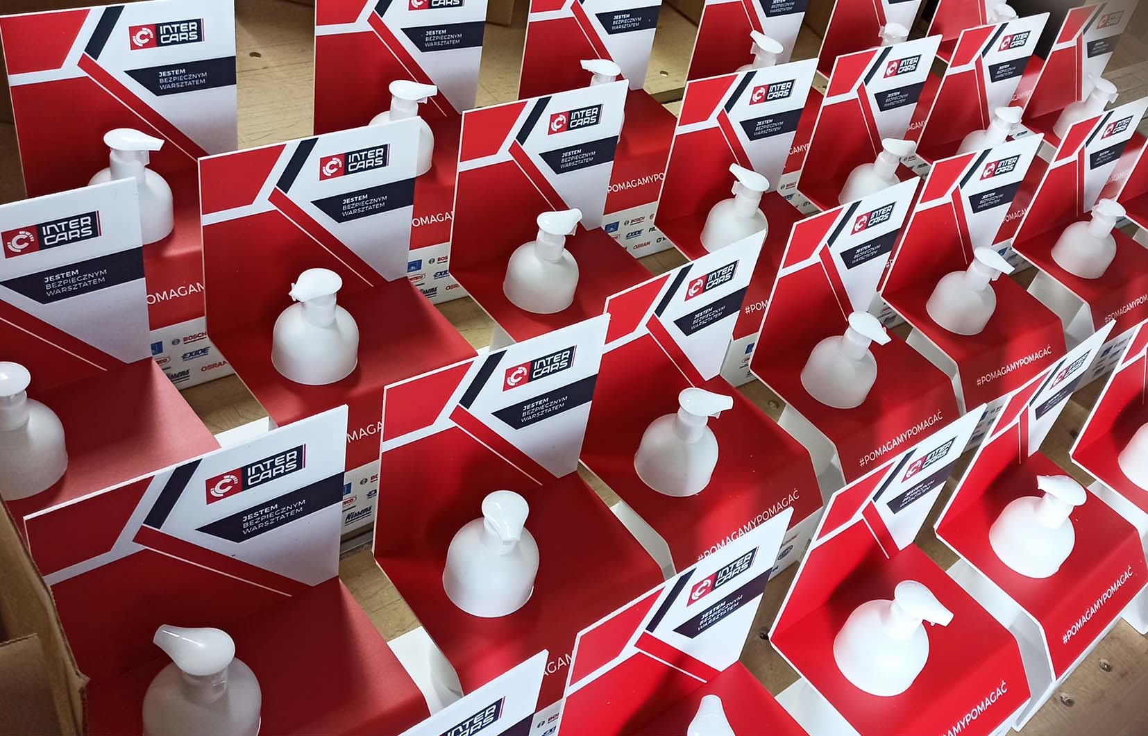 Rząd identycznych zestawów promocyjnych z logo Inter Cars, zawierających biały dozownik na płyn i kartonową podstawkę z hasłem 'Jestem bezpiecznym warsztatem' oraz hashtagiem '#POMAGAMYPOMAGAĆ'.