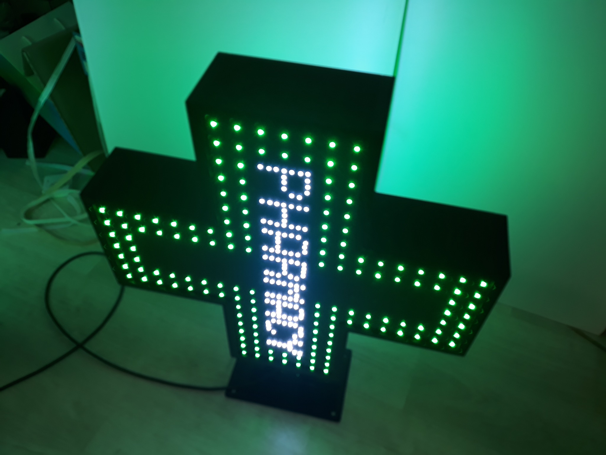 Podświetlany, trójwymiarowy krzyż apteczny LED z napisem 'PHARMA' w pionie, umieszczony na podłodze z zielonym oświetleniem w tle.