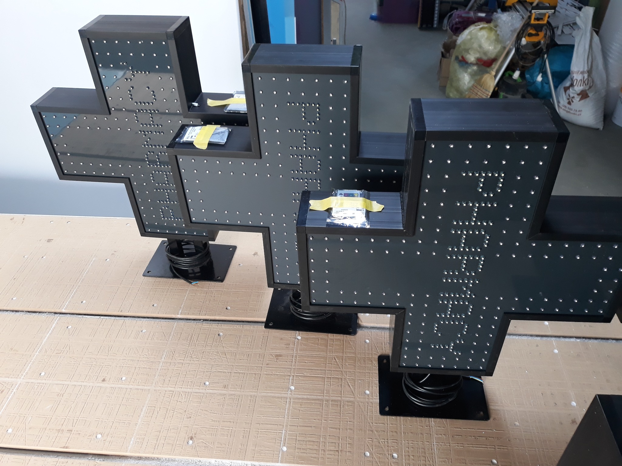 Trzy podświetlane krzyże z napisem 'Apteka' wykonane z czarnego tworzywa, umieszczone na sprężynach, stojące na stole warsztatowym w trakcie montażu.