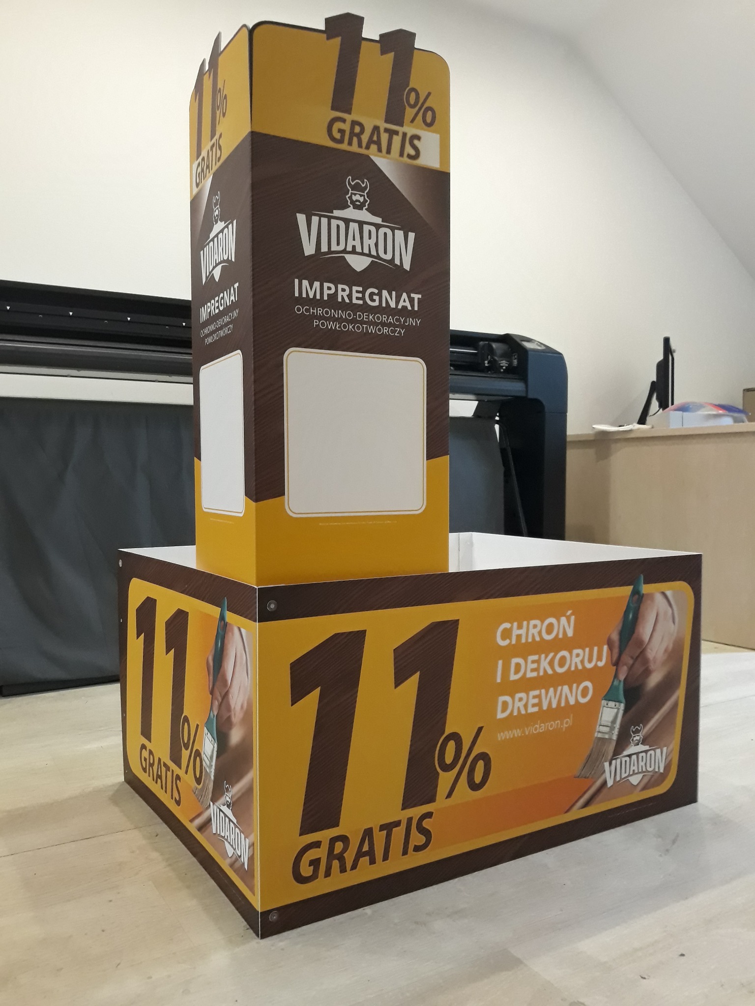 Stojak reklamowy marki Vidaron z hasłem '11% gratis' i informacją o impregnacie ochronno-dekoracyjnym do drewna, w tle ploter tnący.