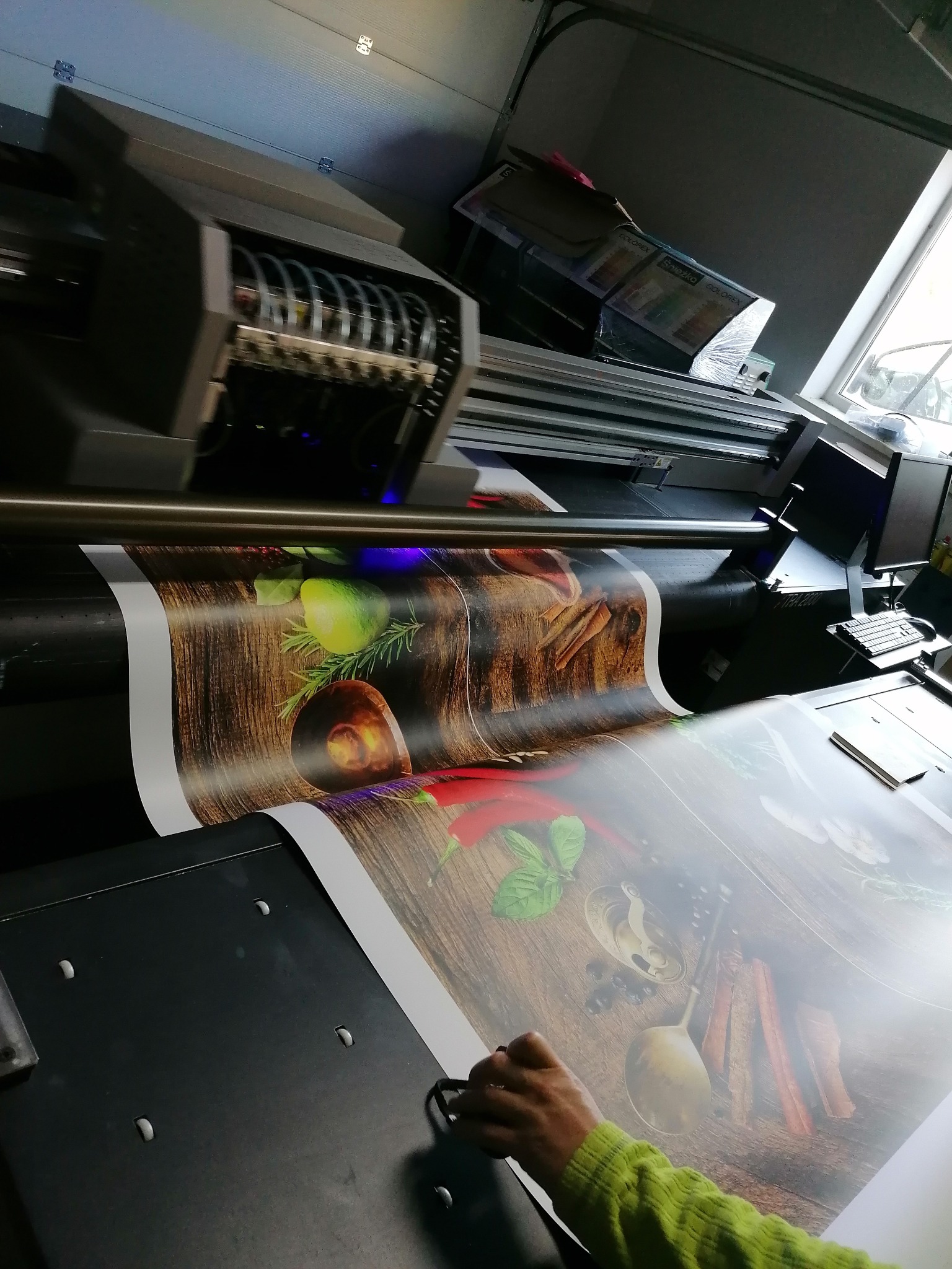Wielkoformatowa drukarka atramentowa w trakcie drukowania grafiki przedstawiającej kompozycję kulinarną z przyprawami i ziołami na drewnianym tle.