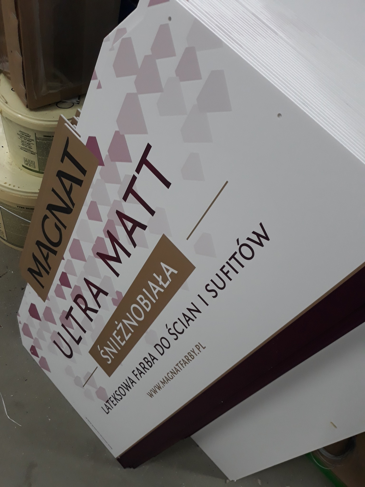 Stos tablic reklamowych farb Magnat Ultra Matt Śnieżnobiała, widoczne opakowania farb w tle.
