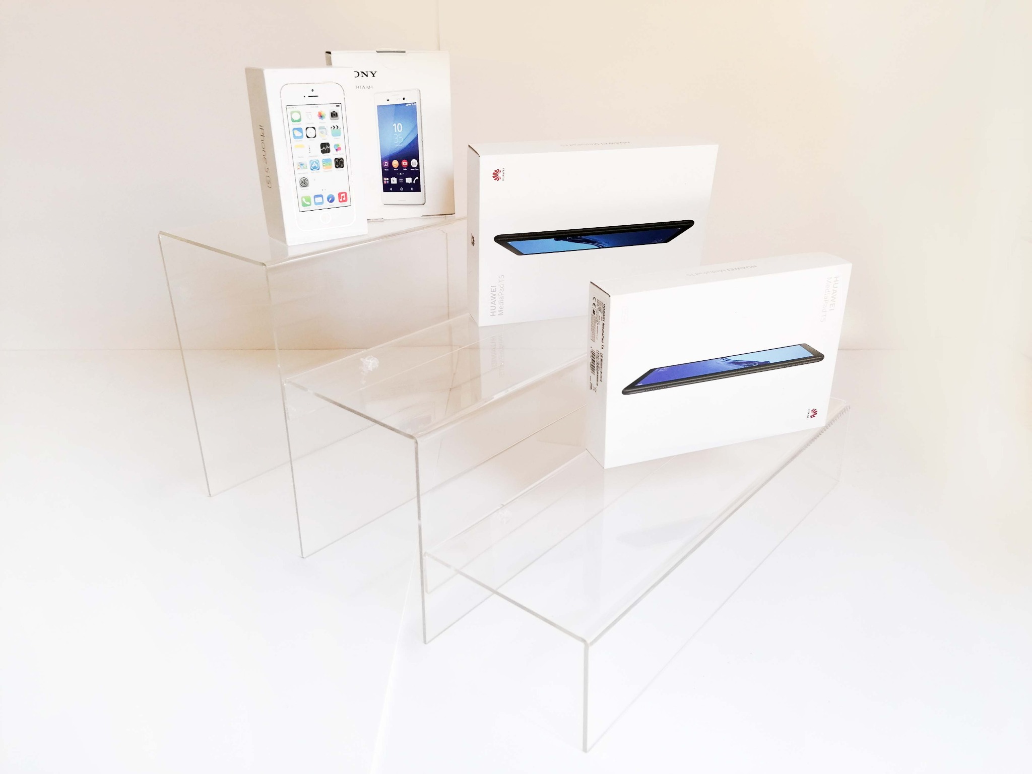 Pudełka z nowymi smartfonami i tabletami (iPhone 5S, Sony Xperia M4, Huawei MediaPad T5) ustawione na przezroczystych, akrylowych schodkach ekspozycyjnych na białym tle.