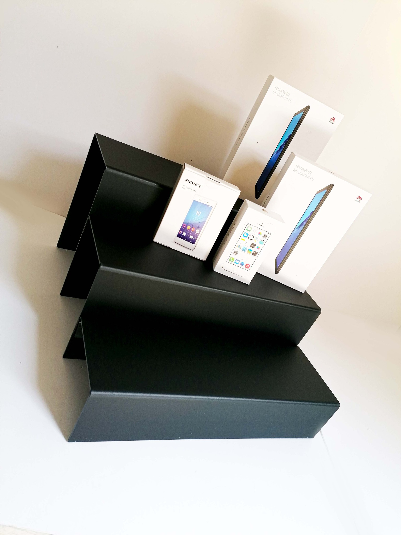 Prezentacja produktów: pudełka smartfonów Sony i atrapa iPhone'a oraz opakowania tabletów Huawei MediaPad T5, ustawione na czarnym, schodkowym stojaku na białym tle.