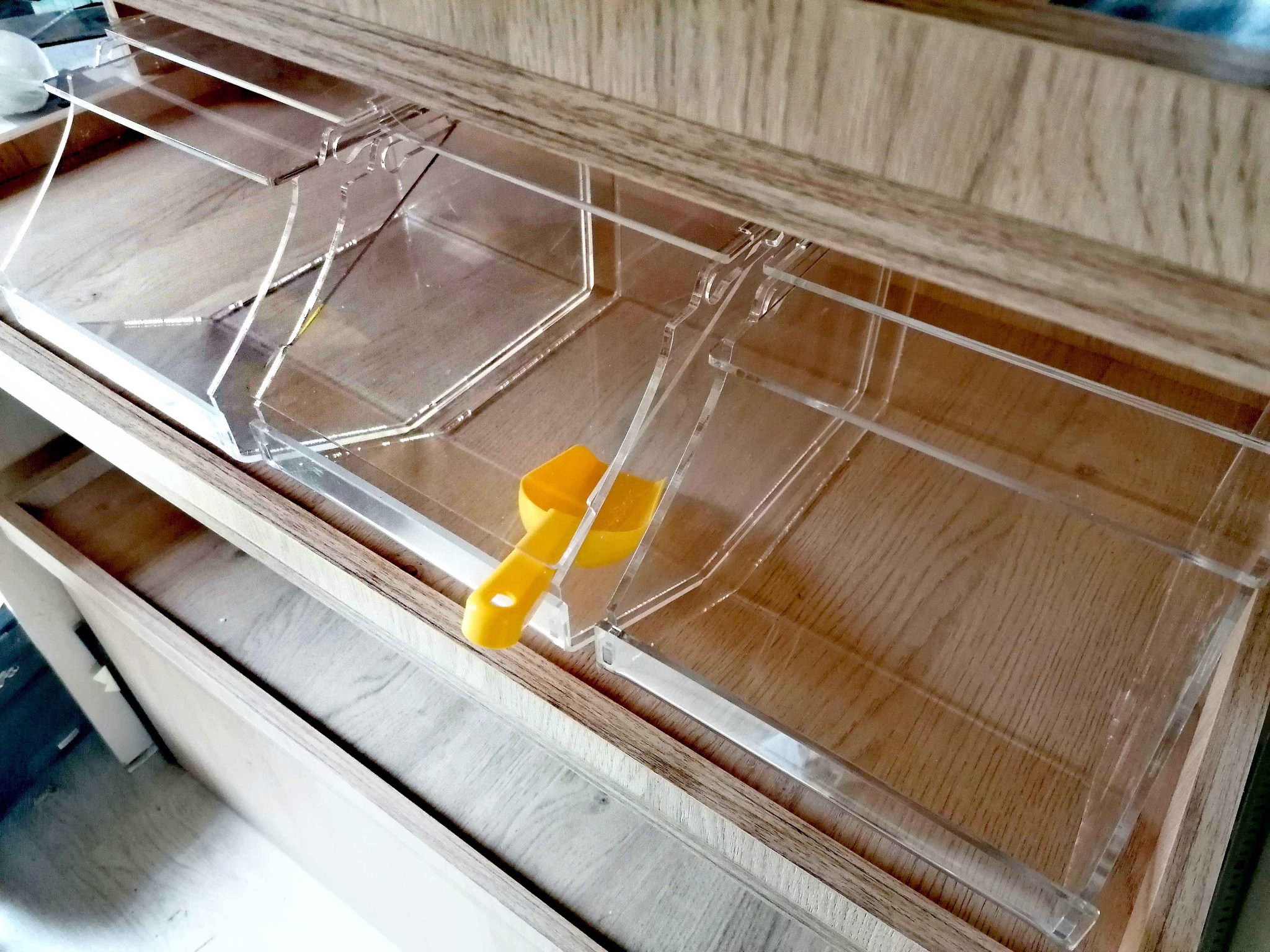 Przezroczyste, akrylowe pojemniki na produkty sypkie w drewnianej zabudowie, z żółtą miarką.