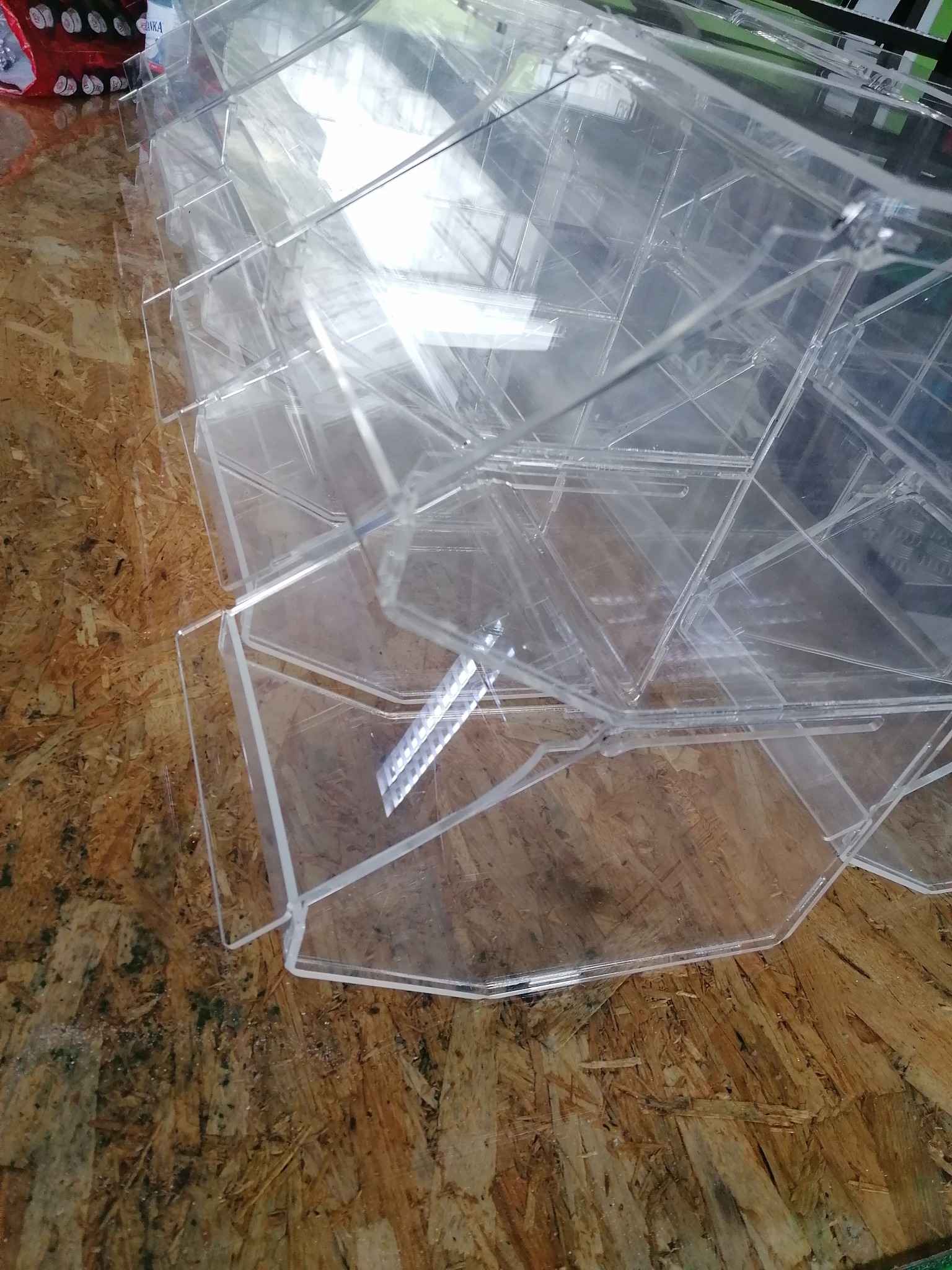 Stos przezroczystych, akrylowych pojemników ekspozycyjnych na blacie z płyty OSB, widoczne niedoskonałości materiału i montażu.