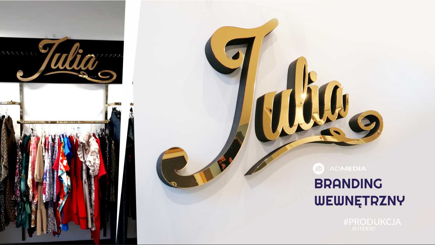 Złoty, przestrzenny napis 'Julia' z czarnym obrysem, zamontowany na białej ścianie, widoczny również w sklepie odzieżowym nad wieszakami z ubraniami.