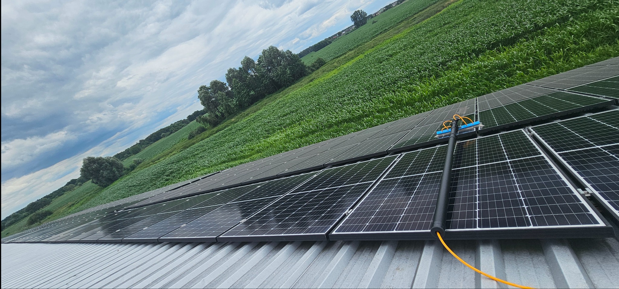 Czyszczenie paneli słonecznych na dachu budynku przy użyciu szczotki teleskopowej z dopływem wody, w tle pole uprawne i pochmurne niebo.