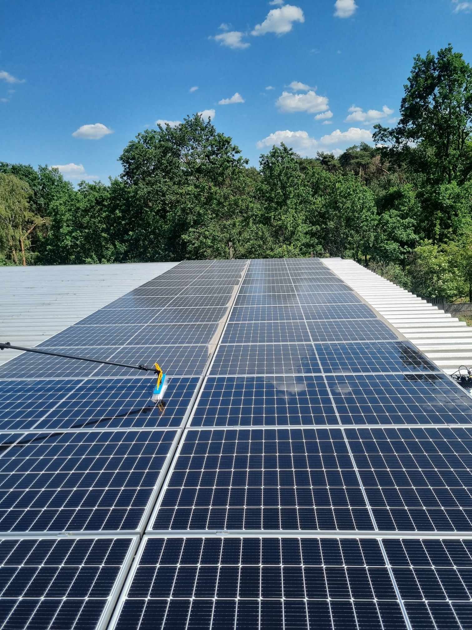 Czyszczenie paneli słonecznych na dachu budynku za pomocą szczotki na długim kiju, widoczne odbicie nieba w panelach.