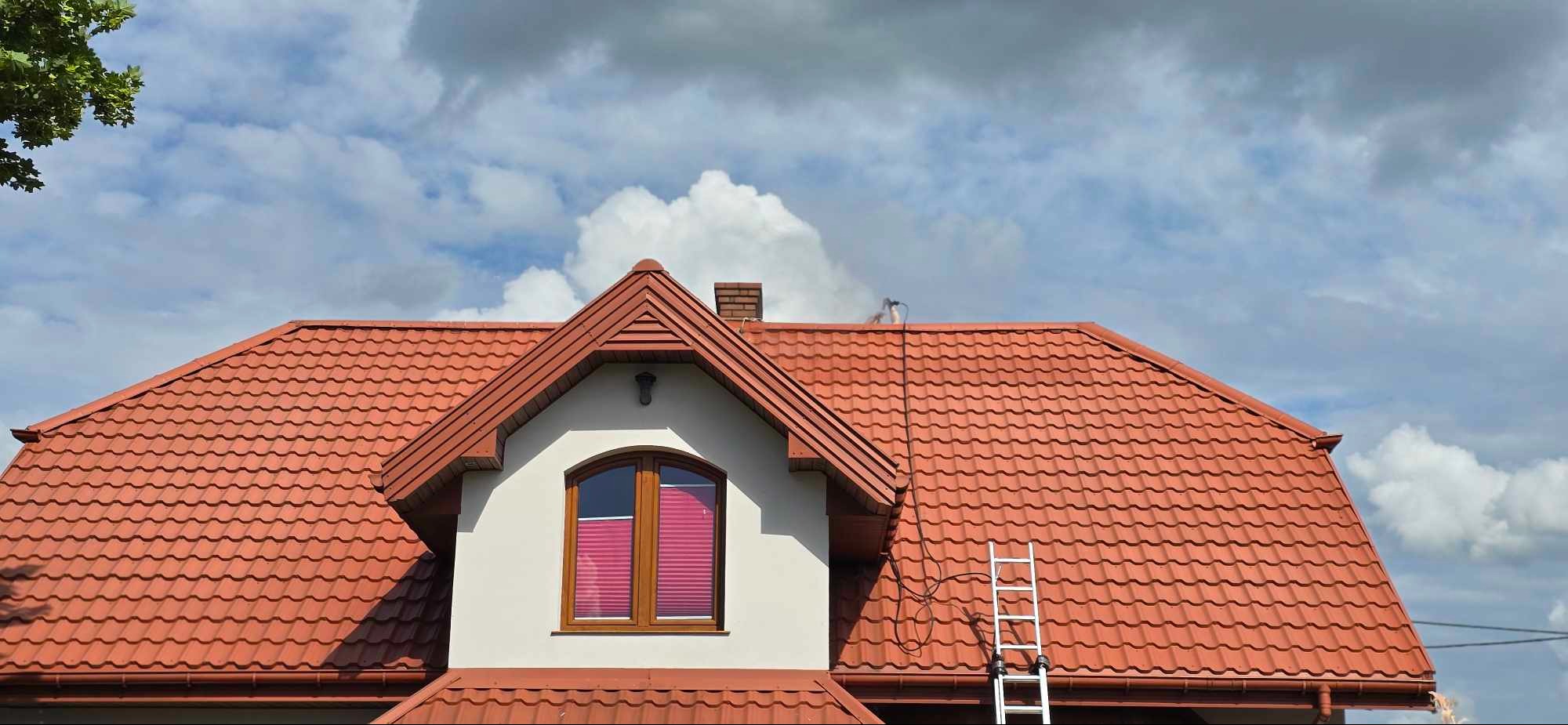 Czerwony dach z dachówki ceramicznej, drabina oparta o dach, widoczne urządzenie do mycia pod ciśnieniem na dachu, błękitne niebo z chmurami w tle.
