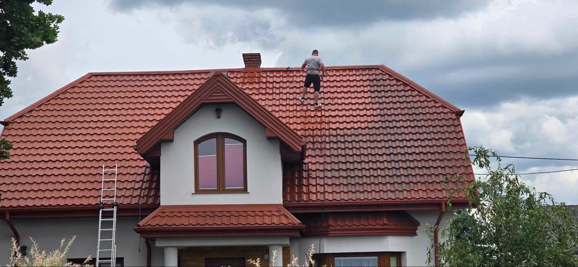 Pracownik zabezpieczony liną asekuracyjną na dachu z czerwonej dachówki podczas prac porządkowych; widoczna drabina oparta o ścianę budynku.