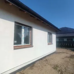 Boczny widok otynkowanego, jasnego domu jednorodzinnego z brązowymi ramami okiennymi, częściowo zabezpieczonymi folią, czarną rynną i odsłoniętym fundamentem, na tle błękitnego nieba i dachu...