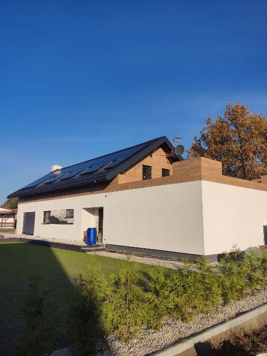 Dom jednorodzinny z częściowo otynkowaną elewacją, widoczne panele słoneczne na dachu i niebieska beczka przy ścianie, trawnik z krzewami na pierwszym planie.