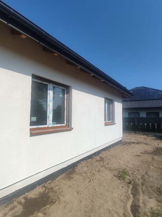 Boczny widok otynkowanego, jasnego domu jednorodzinnego z brązowymi ramami okiennymi, częściowo zabezpieczonymi folią, czarną rynną i odsłoniętym fundamentem, na tle błękitnego nieba i dachu...