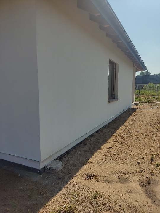 Świeżo otynkowana, biała ściana domu jednorodzinnego z widocznym oknem i fragmentem dachu, otoczona piaszczystym terenem.